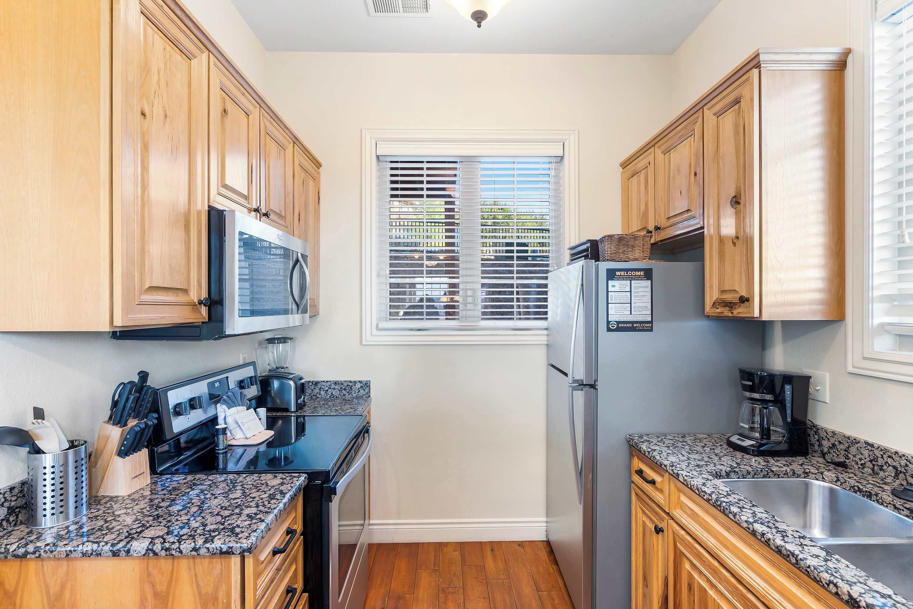 A compact kitchen with wooden cabinets, a black stove, a microwave, a refrigerator, and a window with blinds.
