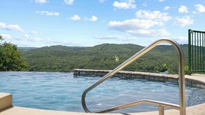 Ein Schwimmbecken mit Metallgeländer, umgeben von einer Betonplattform und Blick auf einen bewaldeten Hügel im Hintergrund.