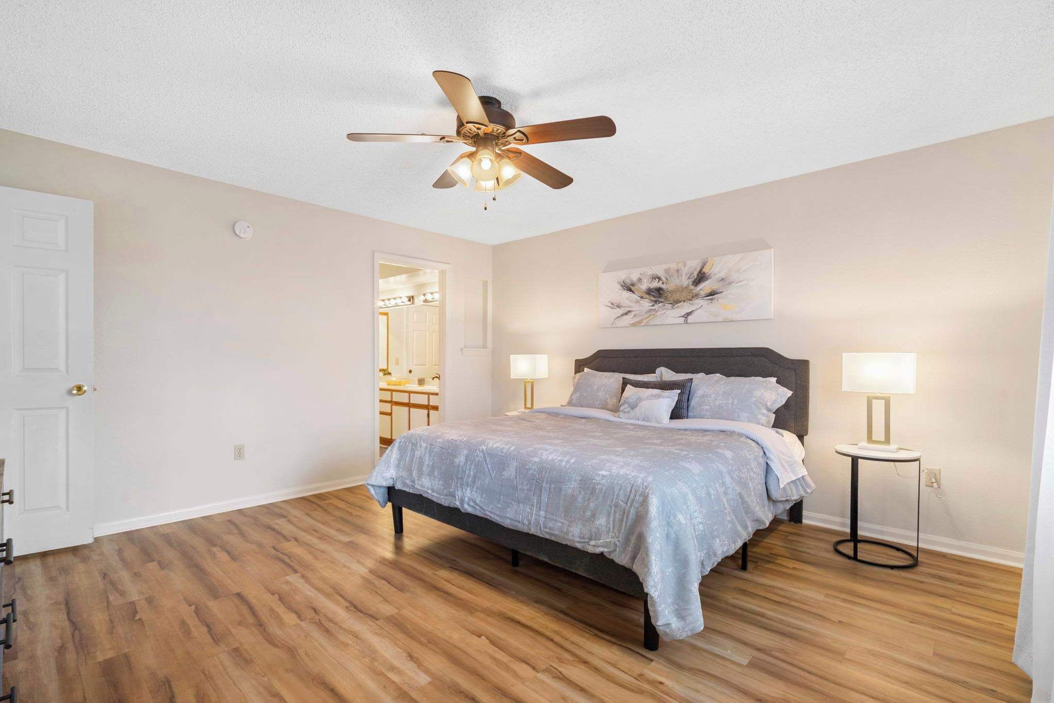 A bedroom with a bed, ceiling fan, bedside lamps, and a painting on the wall.