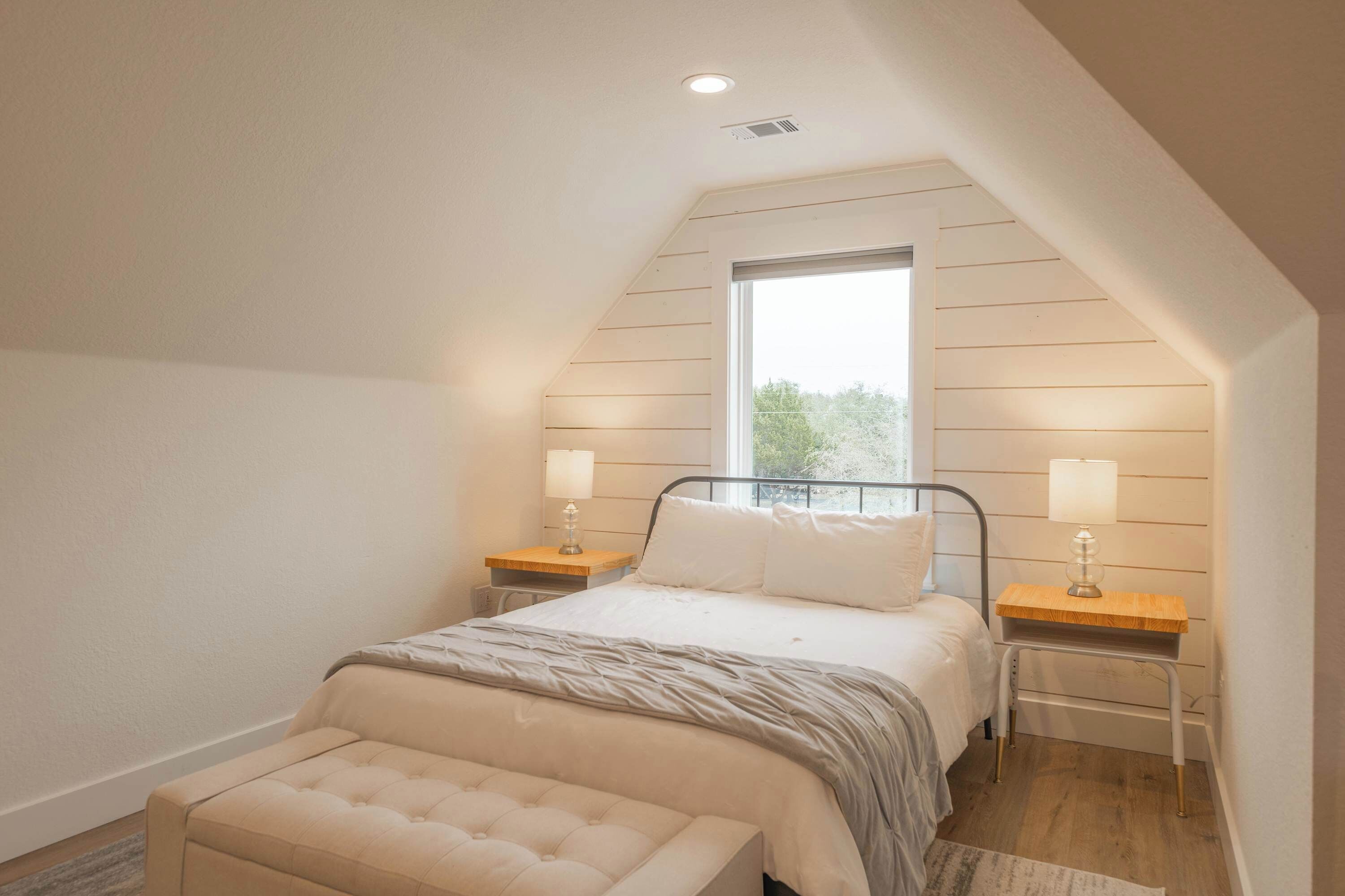 A bedroom with a sloped ceiling, a large window, a bed with white bedding, two bedside tables with lamps, and a bench at the foot of the bed.