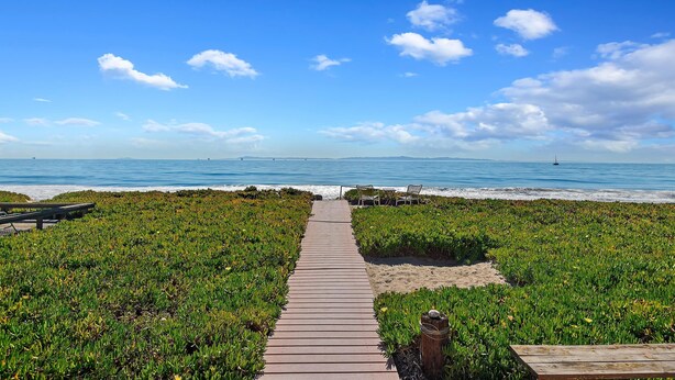 A wooden boardwalk leading to a beach with a clear view of the ocean and a sandy area with greenery on either side.
