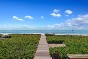 Une promenade en bois mène à une plage d’où l’on a une vue dégagée sur l’océan, et de chaque côté, une zone sablonneuse est bordée de végétation.