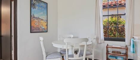 A dining area with a white table and chairs, a poster of a beach, and a window with curtains.