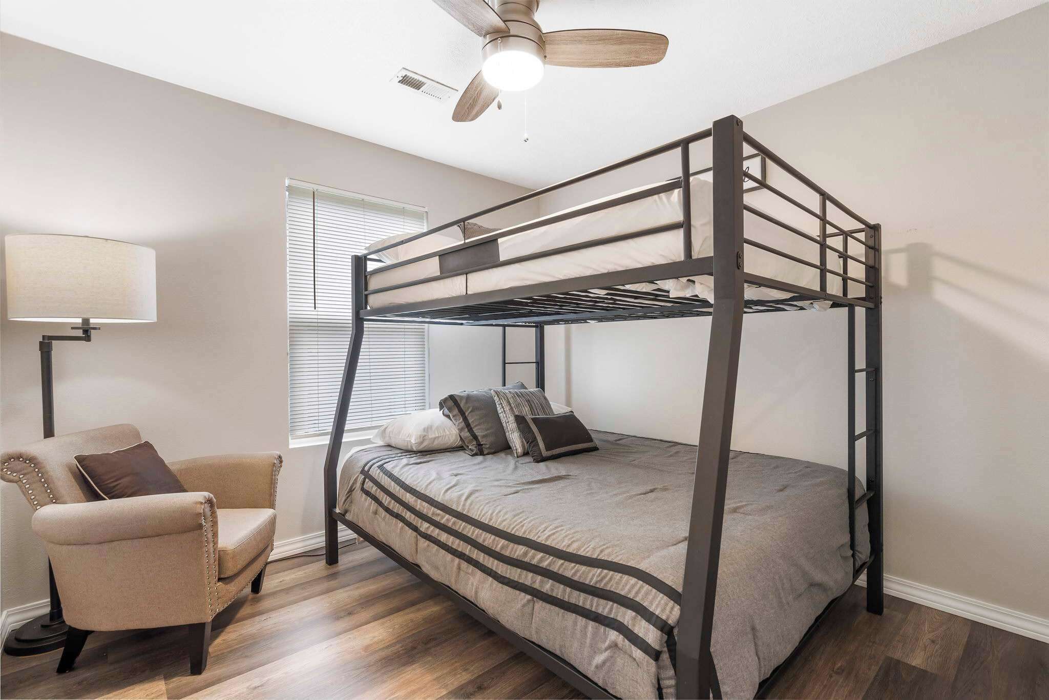 A bunk bed with a ladder, a chair, a lamp, and a ceiling fan in a room with wooden flooring and a window with blinds.