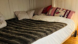 Un lit recouvert d’une couette blanche, d’un plaid épais et sombre, et agrémenté de plusieurs oreillers. La pièce est dotée d’un plancher en bois et de murs blancs.