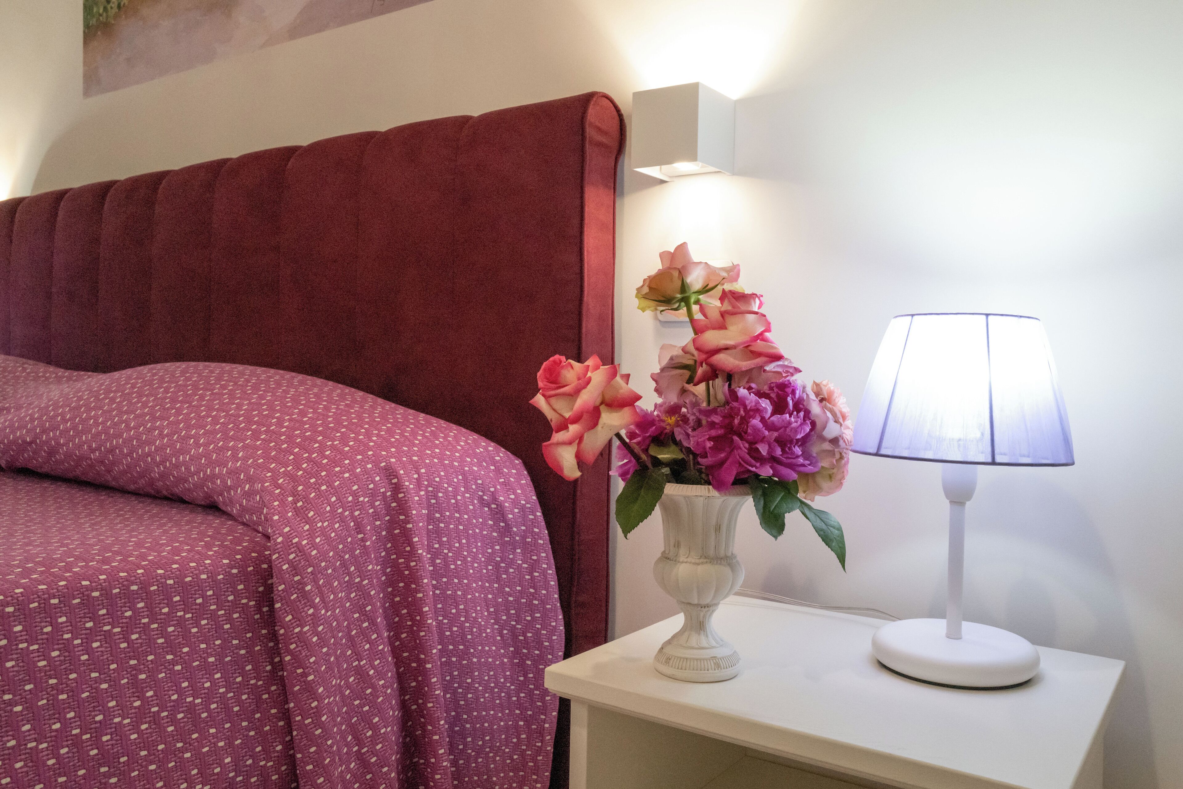 Un lit avec une tête de lit rouge, une table de chevet blanche avec une lampe et un vase de fleurs.