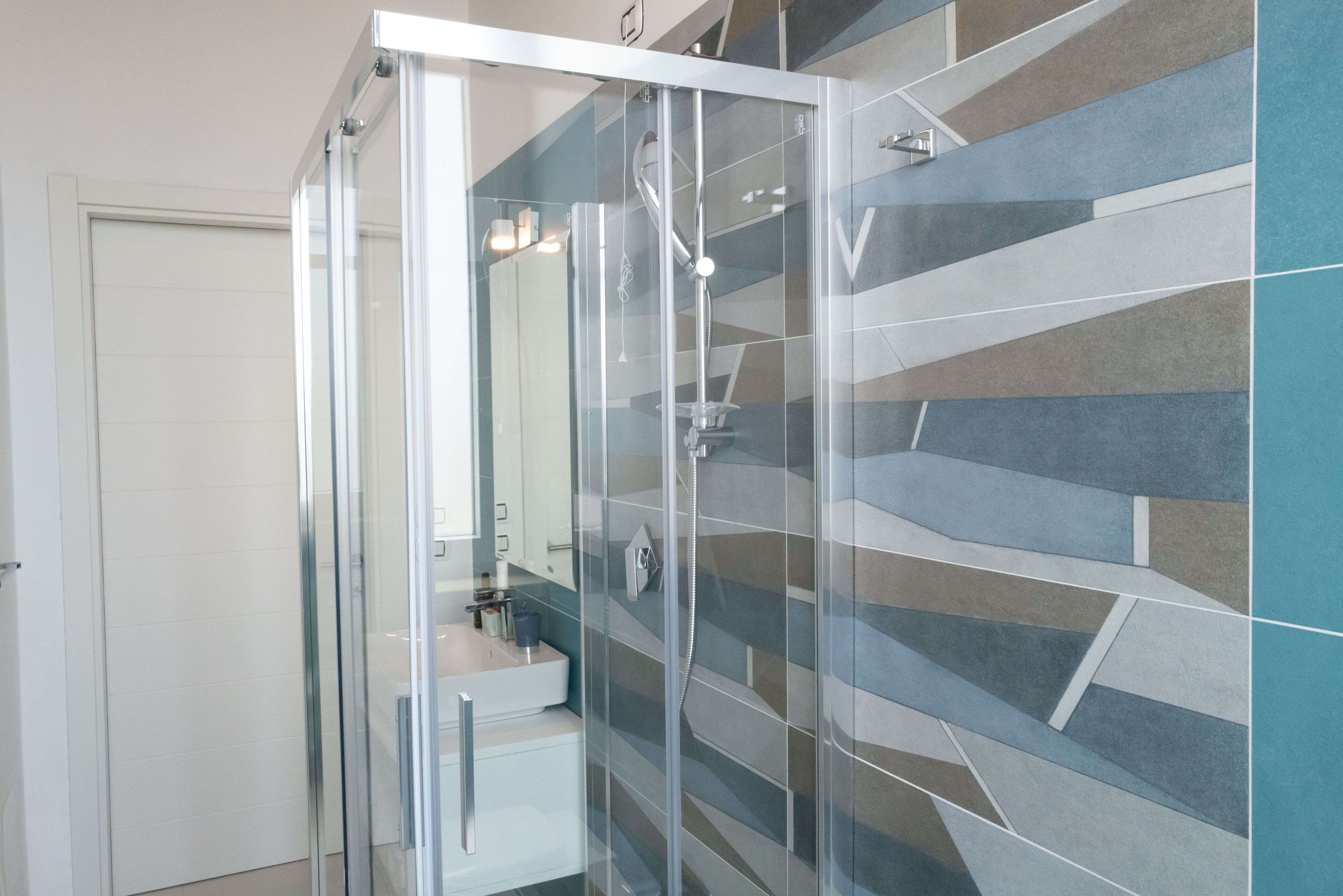 Un baño moderno con ducha envuelta en vidrio, un lavabo blanco y un salpicadero de azulejos geométricos.