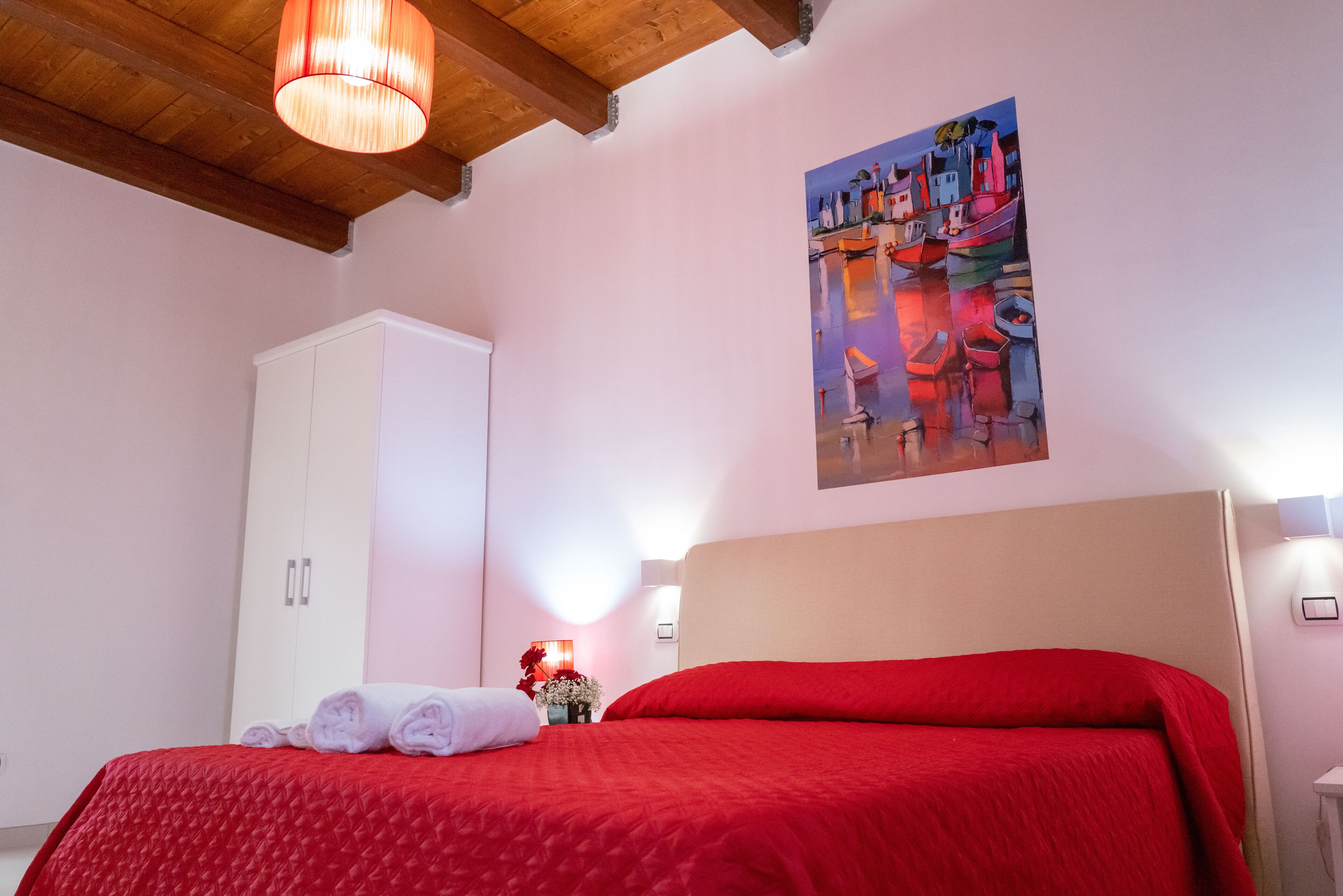 Un dormitorio con una colcha roja, un armario blanco, un cuadro de barcos en la pared y techo de madera.