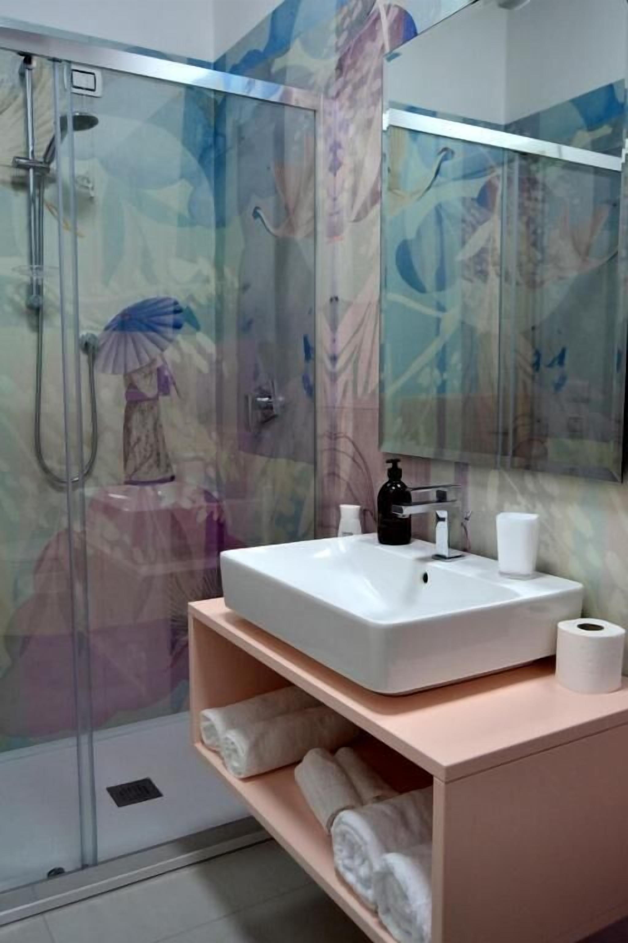 Ein Badezimmer mit Duschkabine aus Glas, einem weißen Waschbecken, einem Spiegel und einem Ablagebereich mit Handtüchern.
