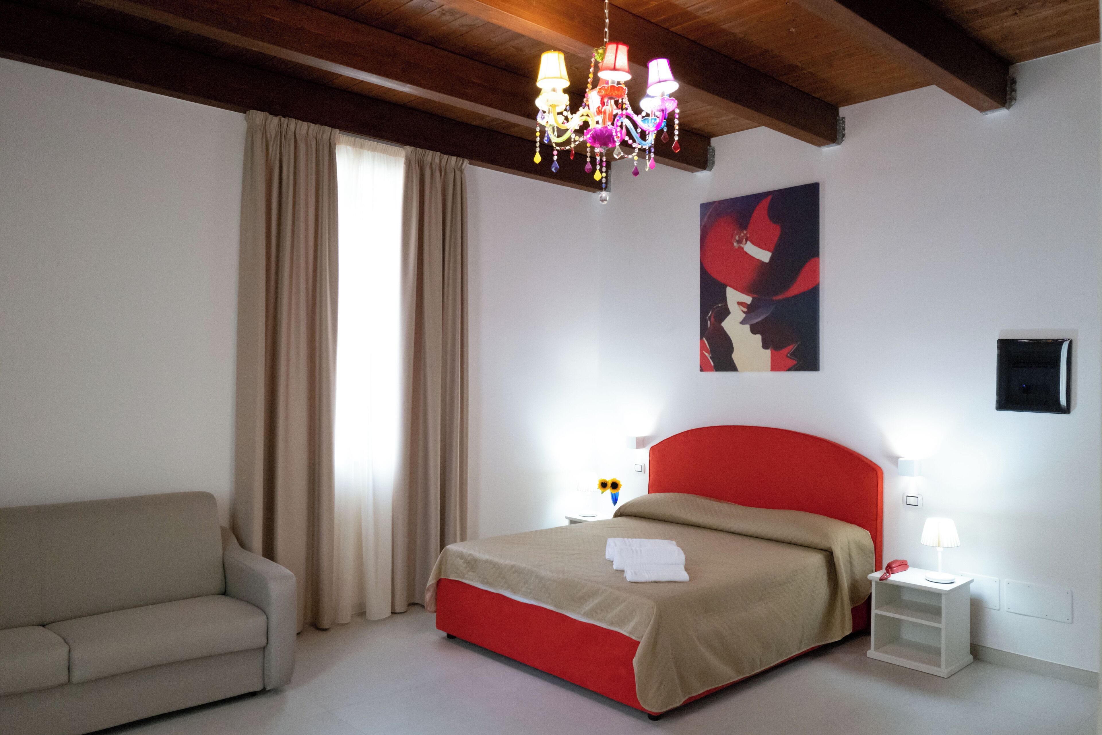 A bedroom with a red bed, beige curtains, a beige sofa, a chandelier, and a painting on the wall.
