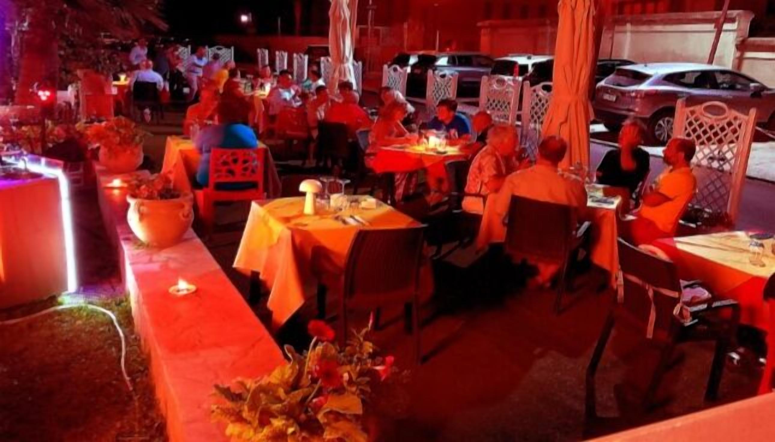Un área de comedor al aire libre con mesas y sillas, algunas con sombrillas. Se ven coches estacionados al fondo.