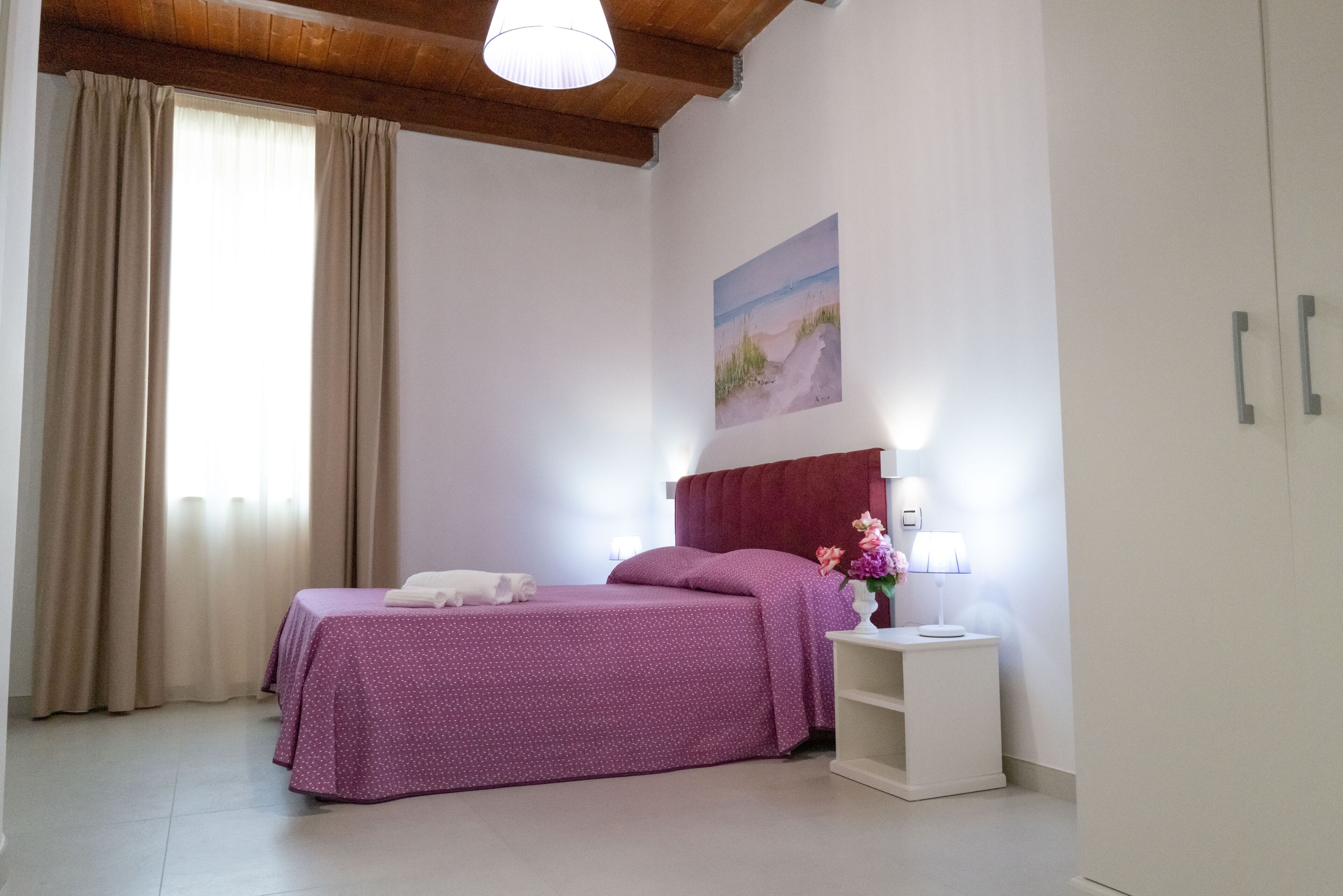 A bedroom with a bed, a nightstand, a lamp, a painting, and a window with curtains.
