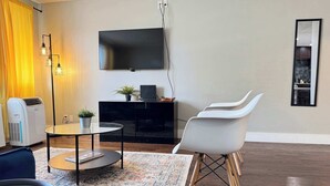 Ein modernes Wohnzimmer mit Fernseher, einem runden Glastisch, einer Topfpflanze, einem weißen Sessel und einem großen Teppich.