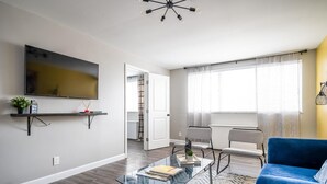 Een moderne woonkamer met een glazen salontafel, een blauwe bank, een wandgemonteerde televisie en een raam met dunne gordijnen.