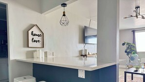 Une cuisine moderne avec un plan de travail blanc, un luminaire suspendu et un panneau décoratif portant l’inscription « Là où il y a de l’amour, là est le foyer ».