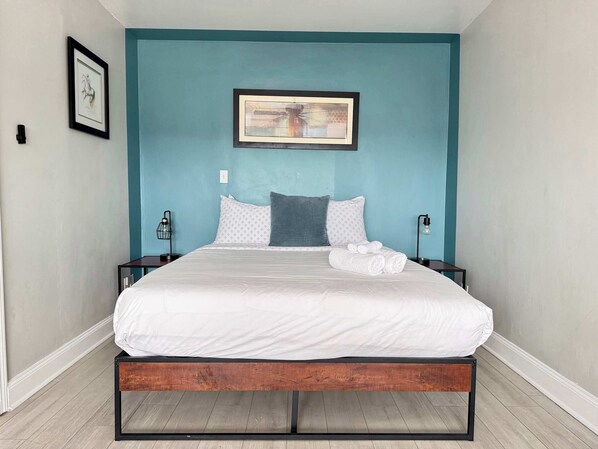 A neatly made bed with white linens, a blue wall, and a framed picture above it.