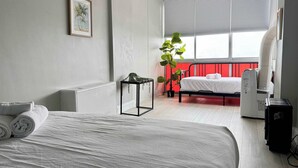 Une chambre d’hôtel avec un lit, un radiateur, un ventilateur de plafond, une fenêtre avec des stores, une plante en pot et un chauffage d’appoint.