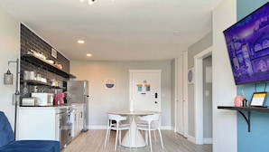 Uma sala de estar moderna com um sofá azul, um espaço de jantar com cadeiras brancas, uma cozinha com frigorífico e micro-ondas, e uma TV fixada na parede.