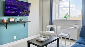 Uma sala de estar moderna com televisão de tela plana na parede, mesa de centro, duas cadeiras brancas, um sofá azul e uma janela com vista para a cidade.