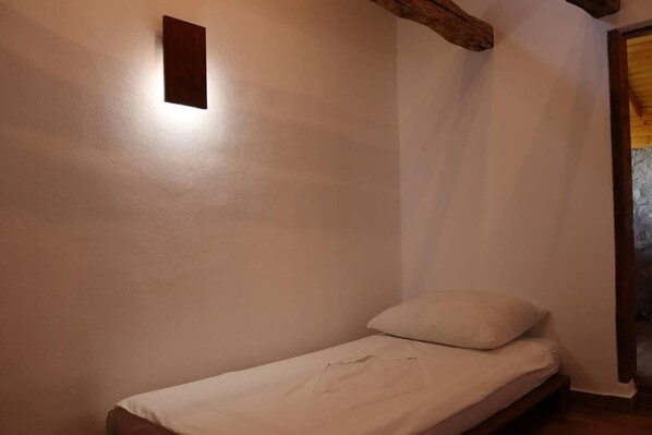Una habitación pequeña con cama individual, cabecera de madera, pared blanca y una lámpara rectangular.