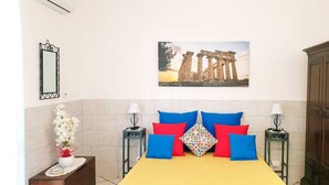 A bedroom with a bed, two bedside tables, a mirror, a vase of flowers, and a wall-mounted picture of ancient ruins. - Il Canneto Vecchia Marina (castelvetrano)