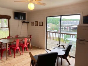 A room with a dining table, chairs, a ceiling fan, a TV mounted on the wall, and a large window with a view of a dock and water.