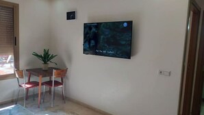 Huoneessa on seinälle asennettu taulutelevisio, pieni pöytä, jossa on kaksi tuolia, ruukkukasvi ja ikkuna, jossa on sälekaihtimet.
