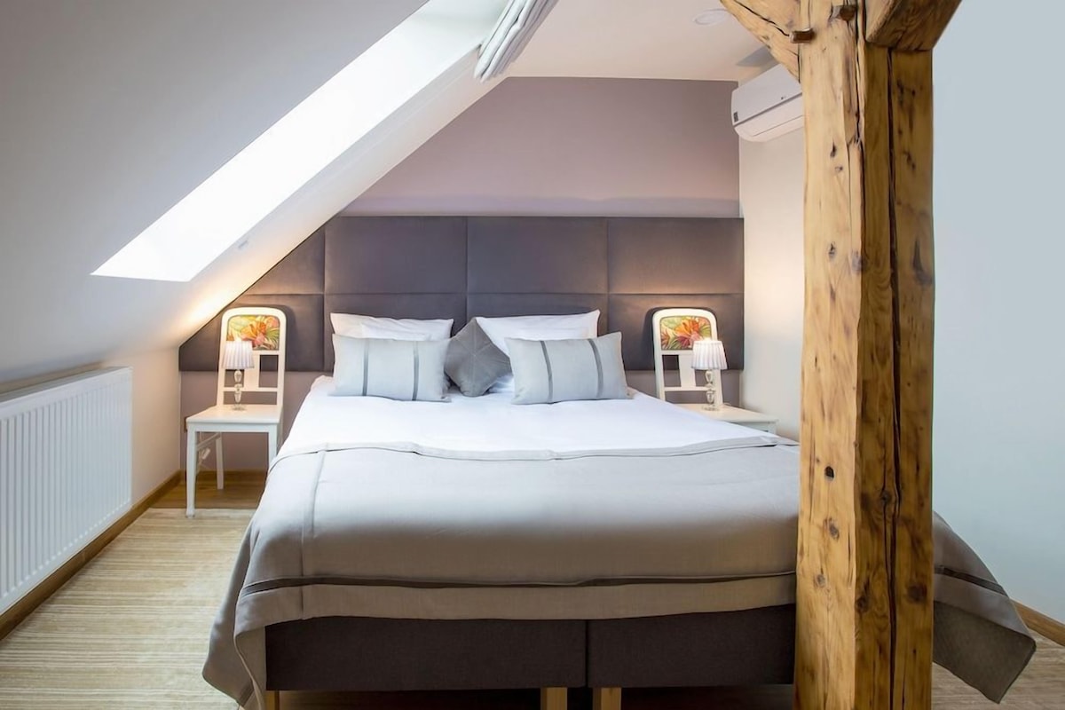 Een slaapkamer met een schuine kap, een bed met veel kussens, nachtkastjes met lampen en een houten balkenstructuur.