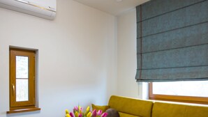 Een woonkamer met een houten salontafel, een gele bank, een raam met zonwering, en een vaas met bloemen.