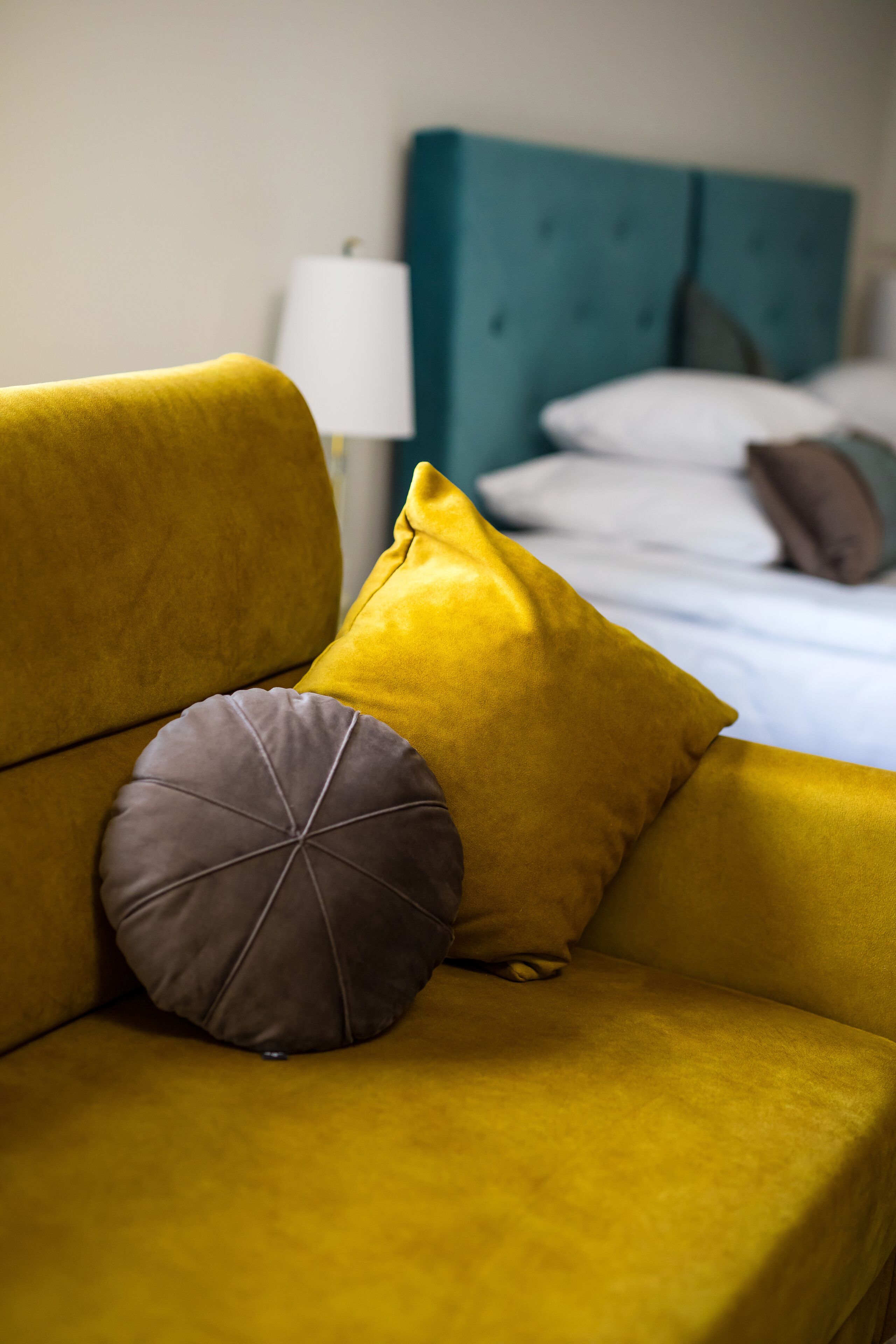 En gul sofa med to puder, en turkis sengegavl og en hvid lampe i et hotelværelse.