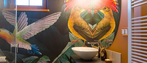 Un baño con un gran mural de pájaros, un espejo redondo, un lavamanos y una ventana.
