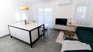 En moderne stue med en grøn sofa, et hvidt sofabord, et fjernsyn og et køkkenområde med en barstol.