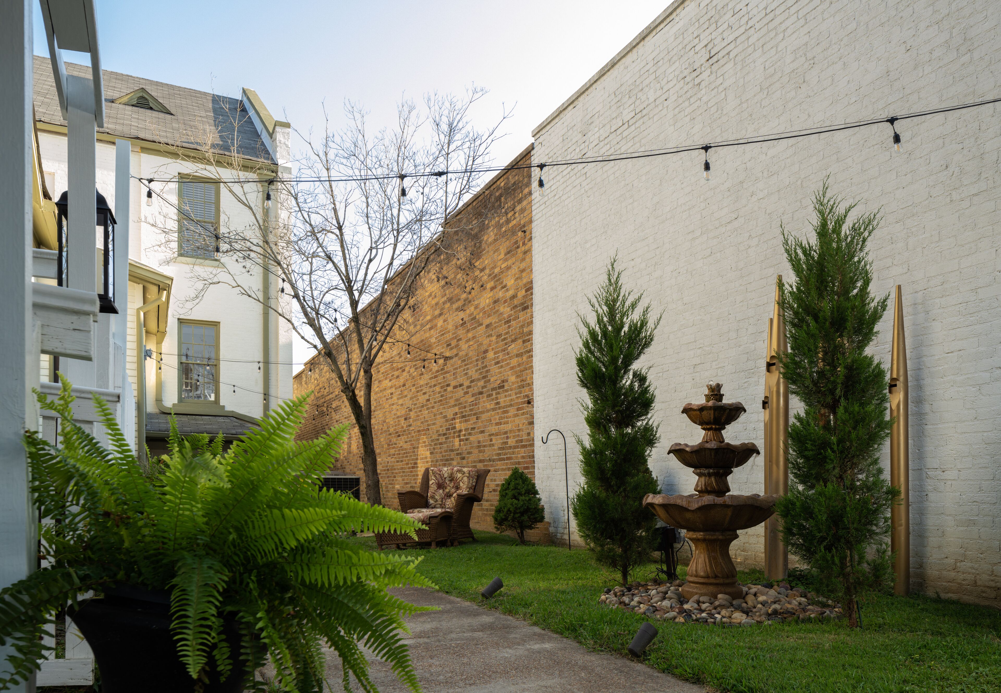Une cour avec un mur de briques, un bâtiment blanc, une fontaine en pierre et une fougère en pot.