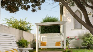 A white wooden bench with yellow cushions, a white pergola with a hanging swing, and a brick wall with greenery. - Magnolia Manor and Chapel (Humboldt)