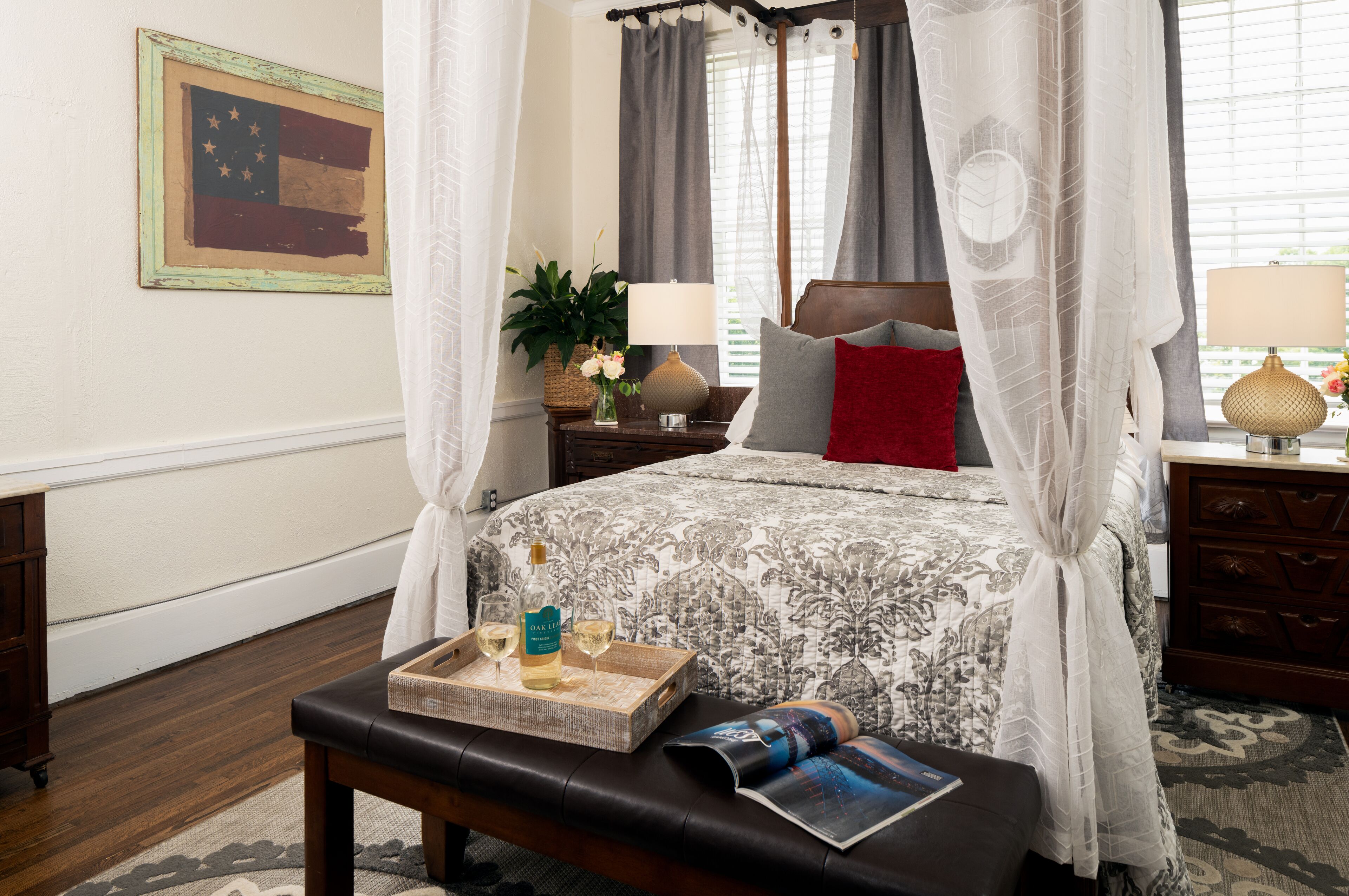 A bedroom with a canopy bed, a bedside table with a lamp, a tray with a bottle and glasses, and a magazine on a bench.