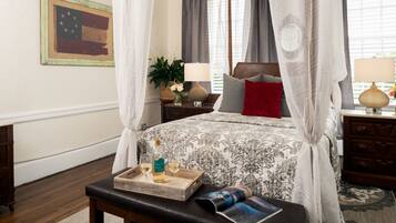 A bedroom with a canopy bed, a bedside table with a lamp, a tray with a bottle and glasses, and a magazine on a bench.