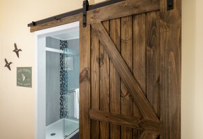 Une porte en bois, de style grange, qui donne accès à une salle de bain dotée d’une douche à parois de verre.
