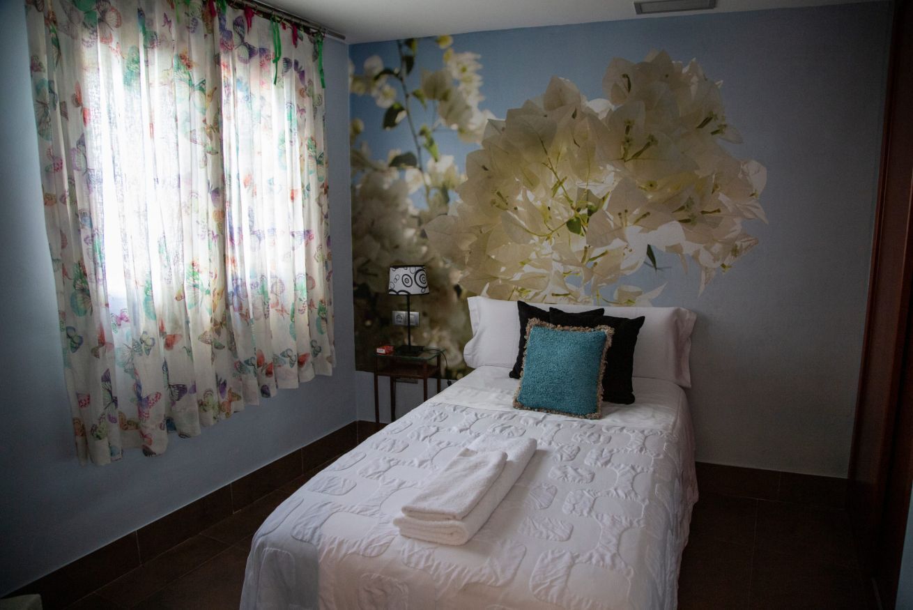 Ein Hotelzimmer mit Bett, einer Wand mit floralem Wandbild, einem Fenster mit Vorhängen und einem kleinen Tisch mit Lampe.