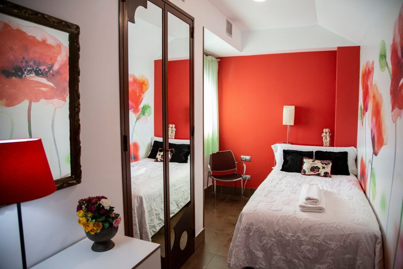 Habitación de hotel con dos camas, un espejo, una pared roja y una florera sobre una mesa.