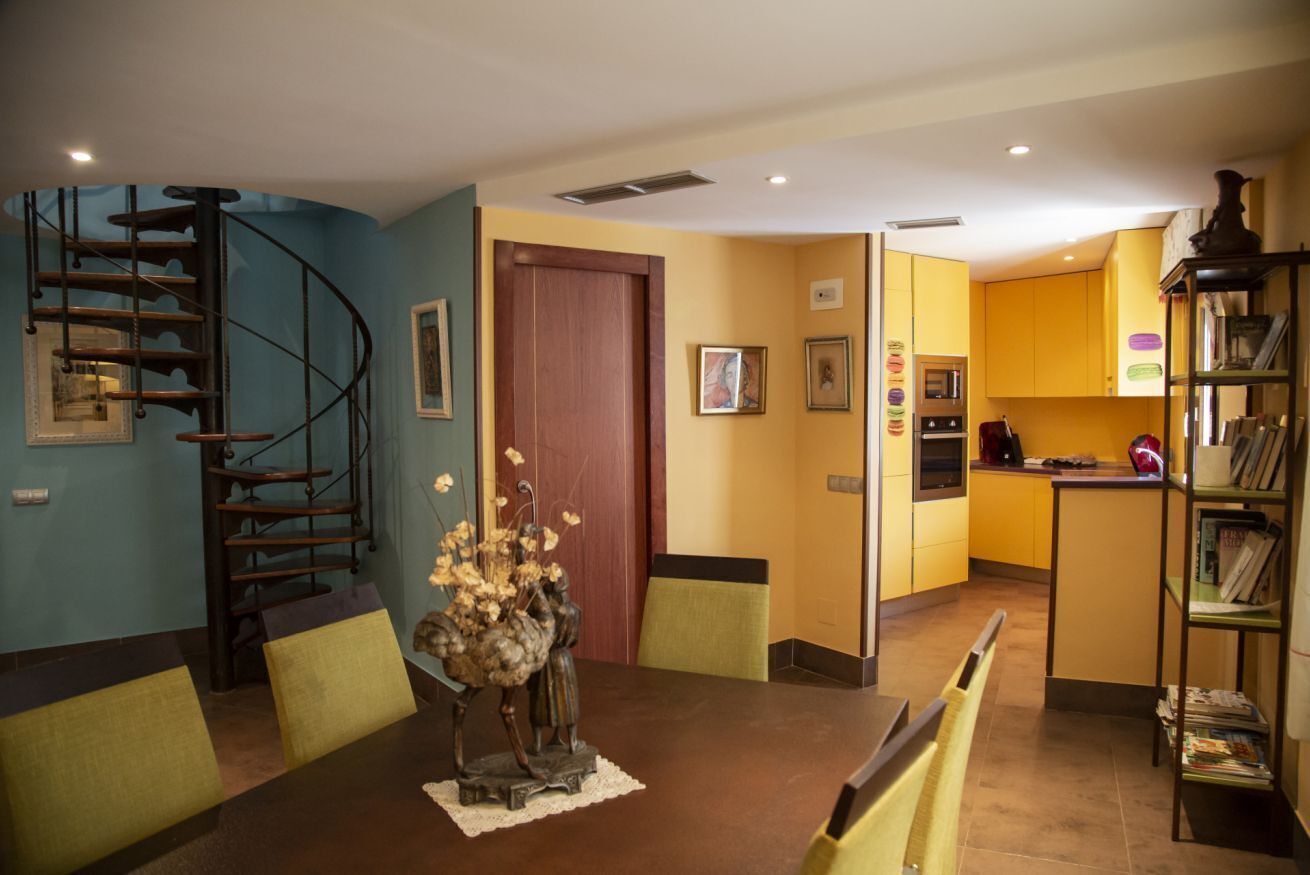 A dining area with a wooden table, chairs, a vase of flowers, a spiral staircase, and a kitchen in the background.