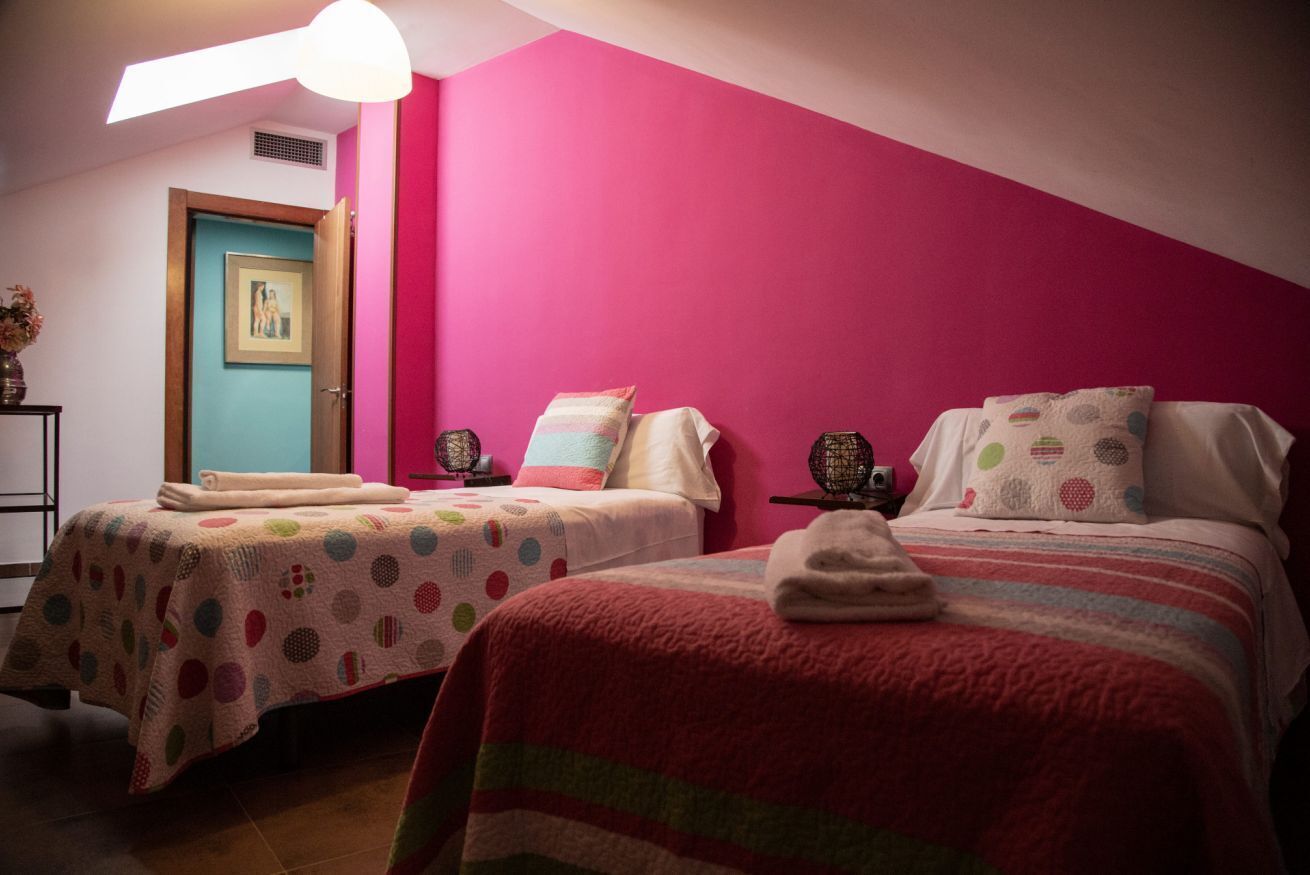 Una stanza con due letti, ognuno con una coperta a fantasia diversa. Si vede una porta con un quadro appeso al muro e un tavolino con un vaso di fiori.
