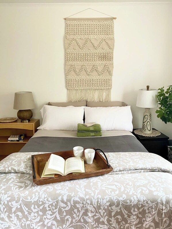 A bedroom with a bed, two bedside tables, a wall hanging, and a tray with a book and two cups.