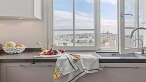 Eine Küche mit Blick auf eine Stadtlandschaft durch ein großes Fenster, inklusive Spüle und einer Arbeitsplatte mit einer Schüssel voller Obst und Gemüse.