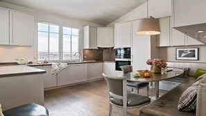 Eine moderne Küche mit weißen Schränken, ein Essbereich mit Tisch und Stühlen sowie ein Blick auf die Stadt durch große Fenster.