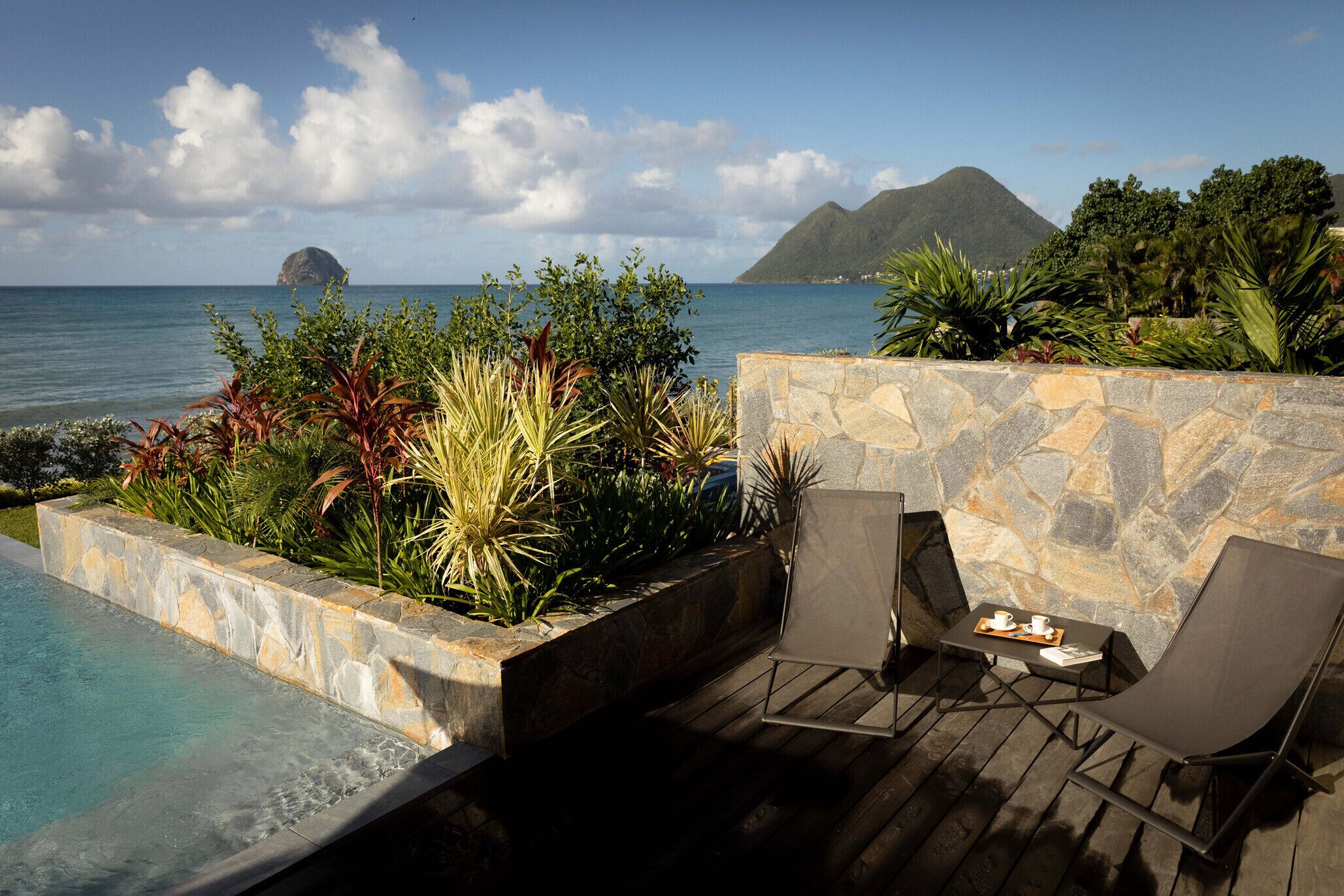 A poolside area with a stone wall, lounge chairs, and a view of the ocean and mountains.