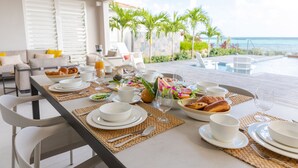 Tavola imbandita per la colazione con piatti, ciotole e tazze bianche, con vista su una piscina e palme.
