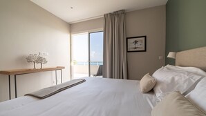 Ein Hotelzimmer mit einem großen Bett, Meerblick durch ein Fenster, einem hölzernen Nachttisch und einem an der Wand befestigten Regal mit Dekoration.