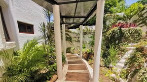 Un sendero techado al aire libre que conduce a un edificio de fachada blanca, rodeado de abundante vegetación.