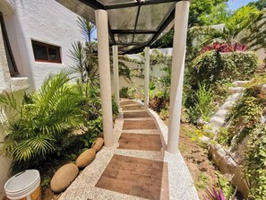 Un percorso coperto conduce a un edificio con facciata bianca, circondato da rigogliosa vegetazione e piante.