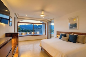 A bedroom with a large bed, a flat-screen TV, a ceiling fan, and a view of mountains through a large window.